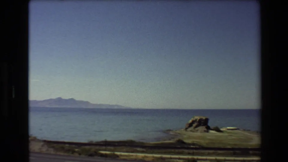 1980 Western USA 8mm frame with a broad sea horizon, distant mountains, clear sky, and a rocky coastal point beside the road.
