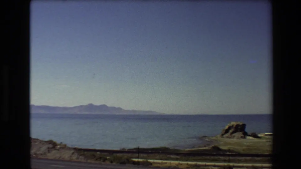 1980 Western USA 8mm frame showing mountains beyond calm water, a low roadside foreground, and a rocky shoreline edge.