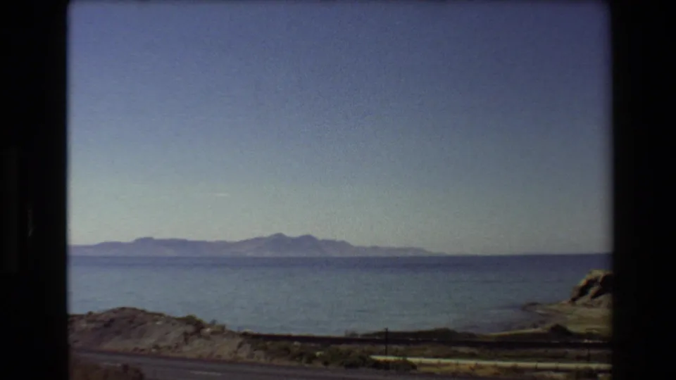 1980 Western USA 8mm frame looking over roadside foreground toward blue water, distant mountains, and a partial rocky shore.