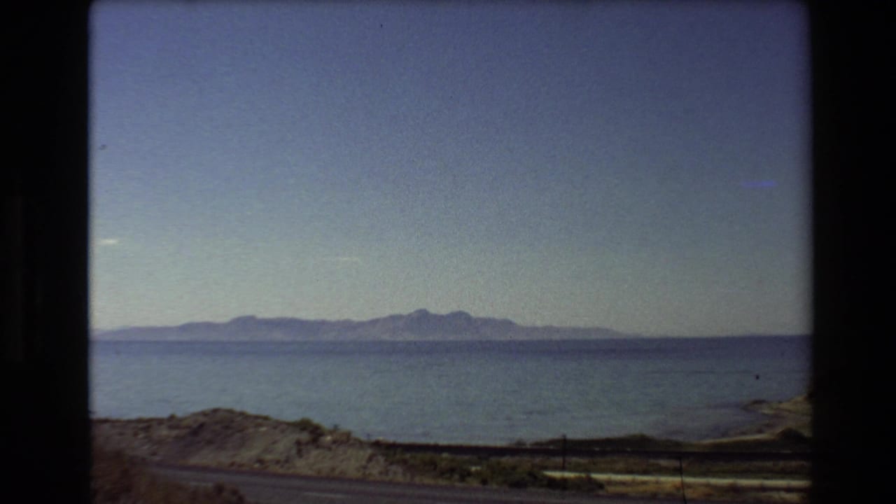 1980 Western U.S. home-movie frame showing a broad freshwater body, low shoreline, distant mountains, and blue sky from a moving vehicle.