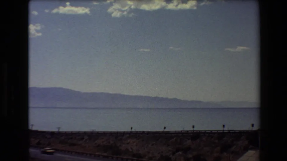 1980 Western U.S. travel footage frame showing a broad lake, distant mountain ridge, sparse clouds, and roadside guardrail from a vehicle.