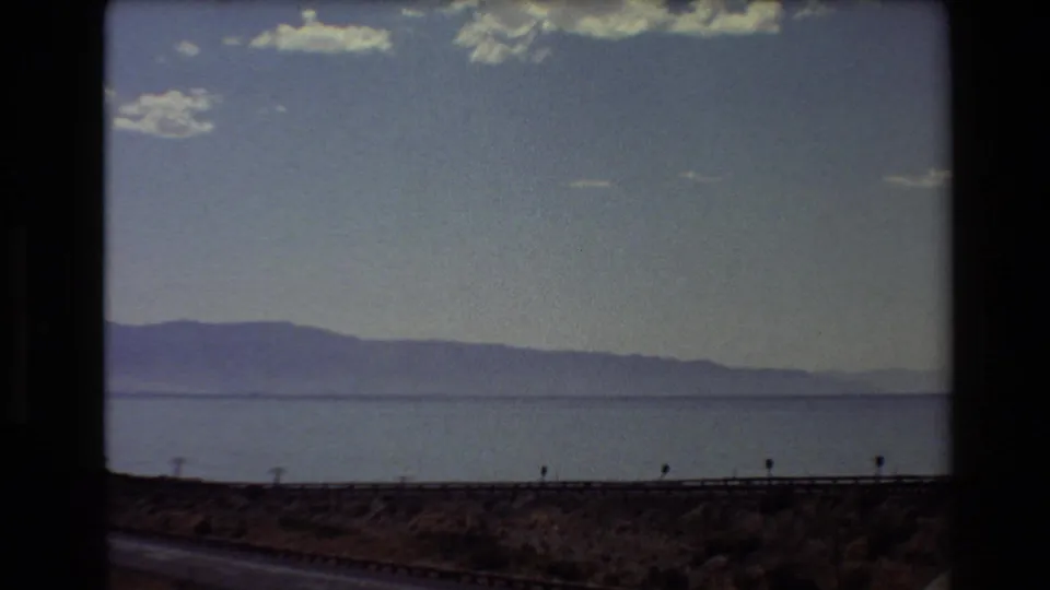 Western U.S. 1980 home-movie frame of a lake and distant mountains, filmed past a roadside guardrail under scattered clouds.