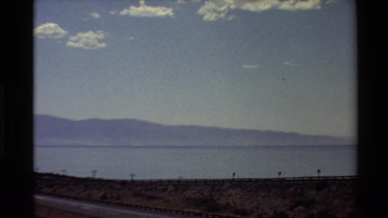 1980 8mm Western U.S. travel view from a moving vehicle, with road, water, mountains, and scattered clouds.