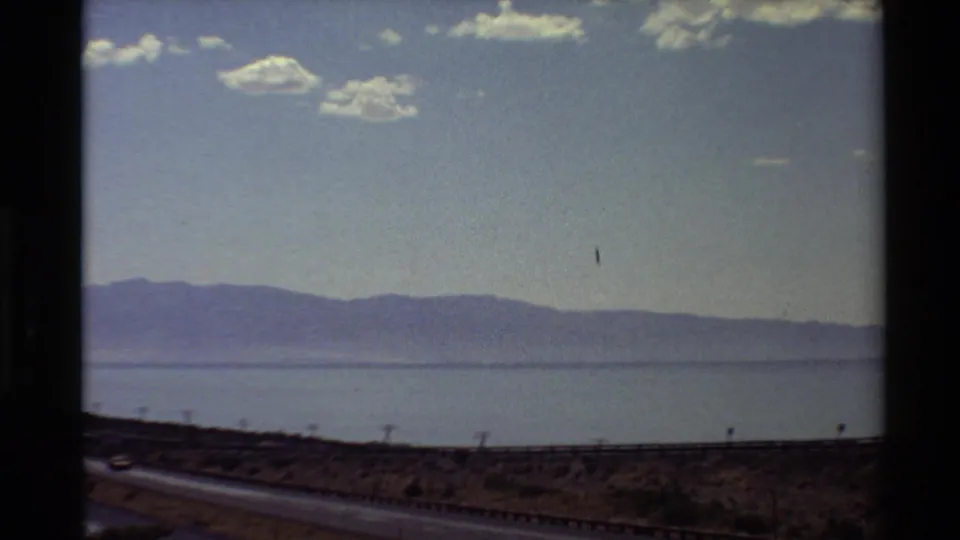 Western U.S. 8mm frame showing a roadside foreground, expansive water, and mountains beneath a partly cloudy sky.