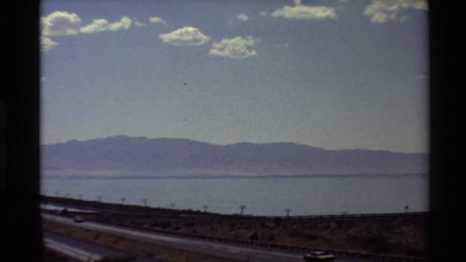 1980 home-movie frame from a moving vehicle, with a passing car, roadside barrier, water, and distant mountains.