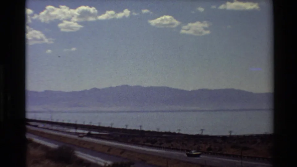 Western U.S. 1980 8mm travel frame showing cars on a shoreline highway with water and mountains beyond.