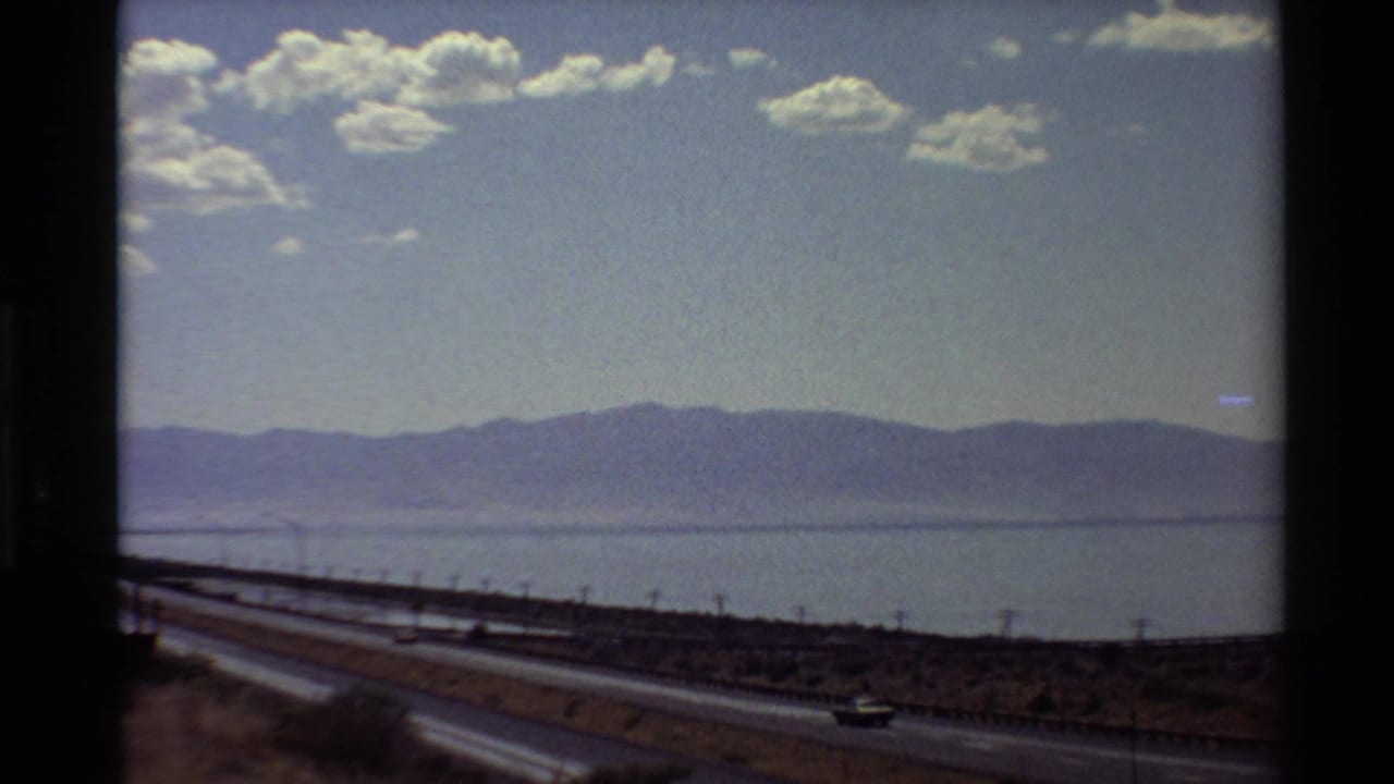1980 Western U.S. home-movie view of a highway running beside broad water, with distant mountains, clouds, and a small car visible.