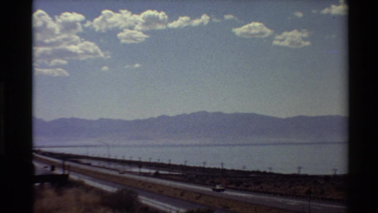 Moving-vehicle 8mm view of a Western U.S. highway beside water, with low mountains and scattered clouds in the distance.