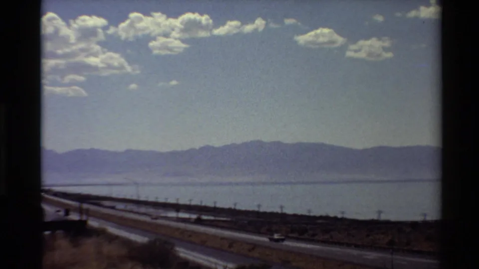 Moving-vehicle 8mm view of a Western U.S. highway beside water, with low mountains and scattered clouds in the distance.