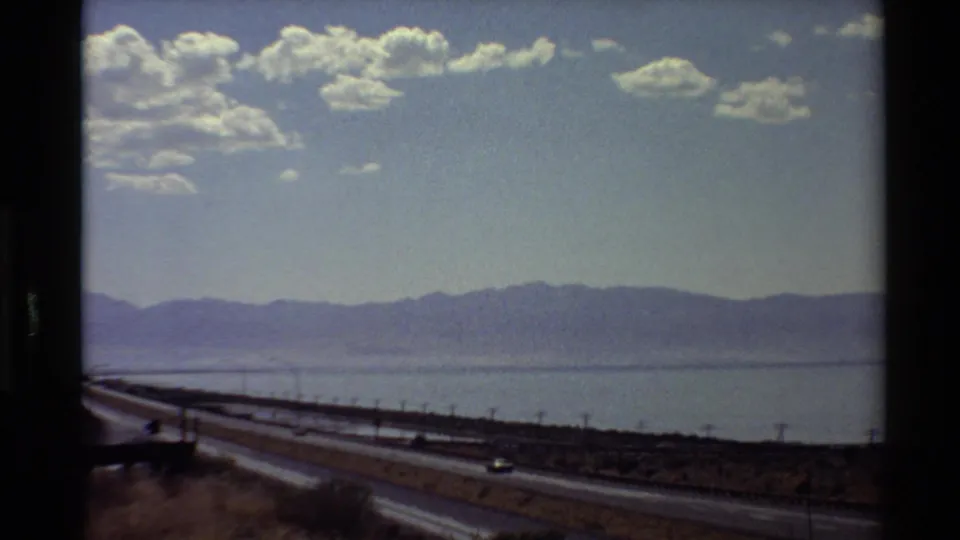 Passenger-view frame of a highway near broad water and mountains, with grain, soft focus, and dark gate edges from the 8mm scan.