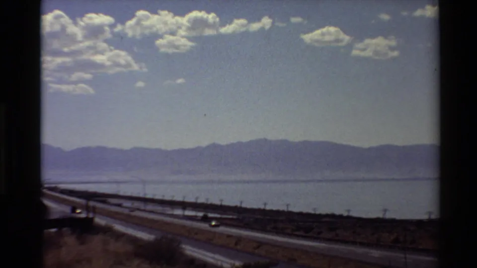 Western U.S. roadside view showing highway lanes, dry foreground land, utility poles, water, and a distant mountain range.