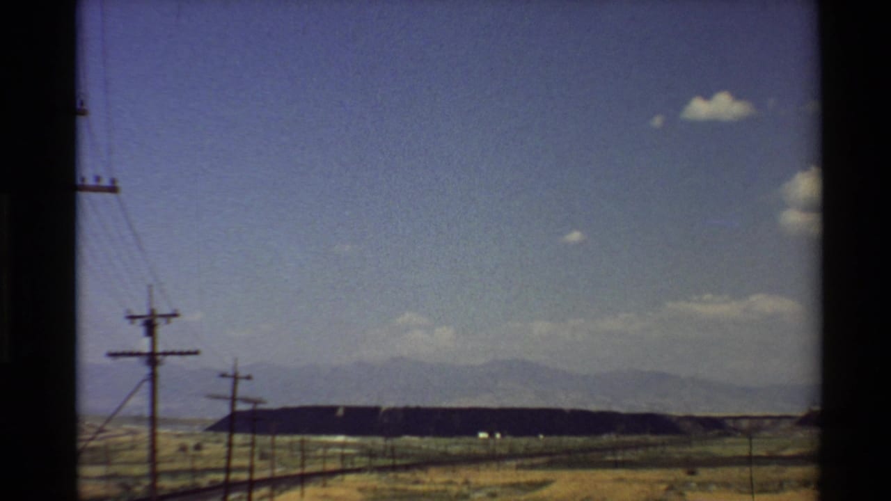 1980 Western U.S. home-movie frame from a moving vehicle showing dry fields, utility poles, and distant mountains under a blue sky.
