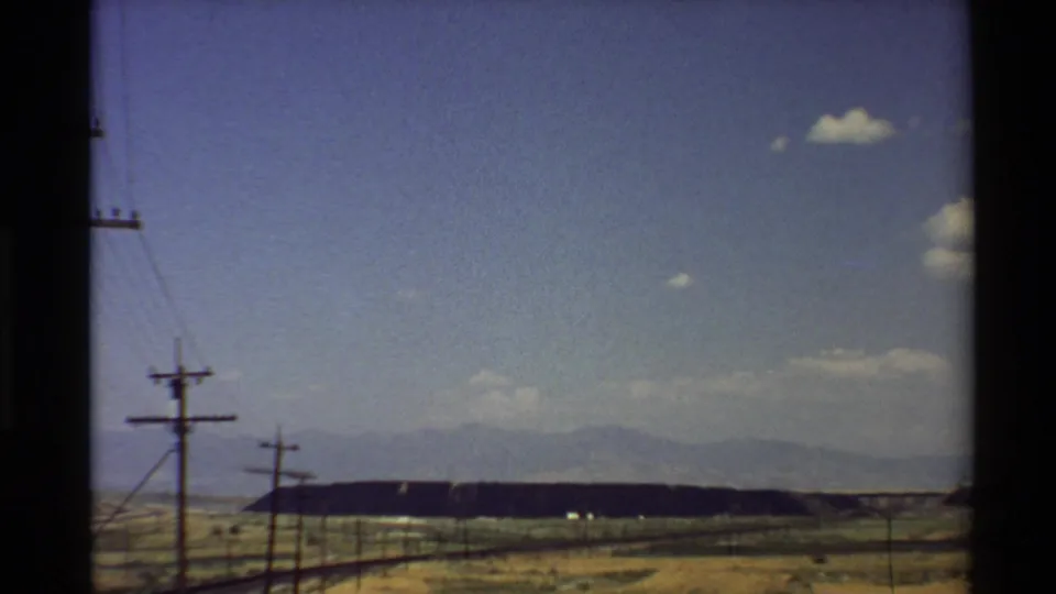 1980 Western U.S. home-movie frame from a moving vehicle showing dry fields, utility poles, and distant mountains under a blue sky.