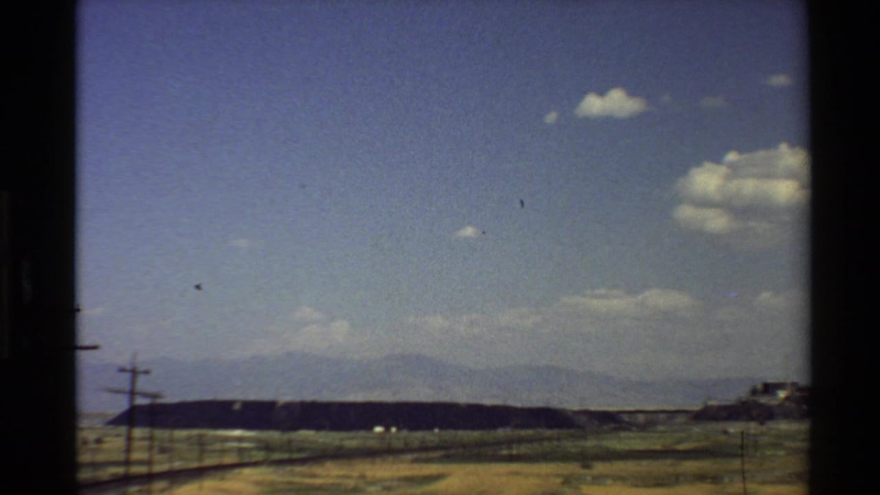 Western U.S. 8mm frame showing an open dry landscape, roadside utility poles, a low ridge, and hazy mountains beyond.