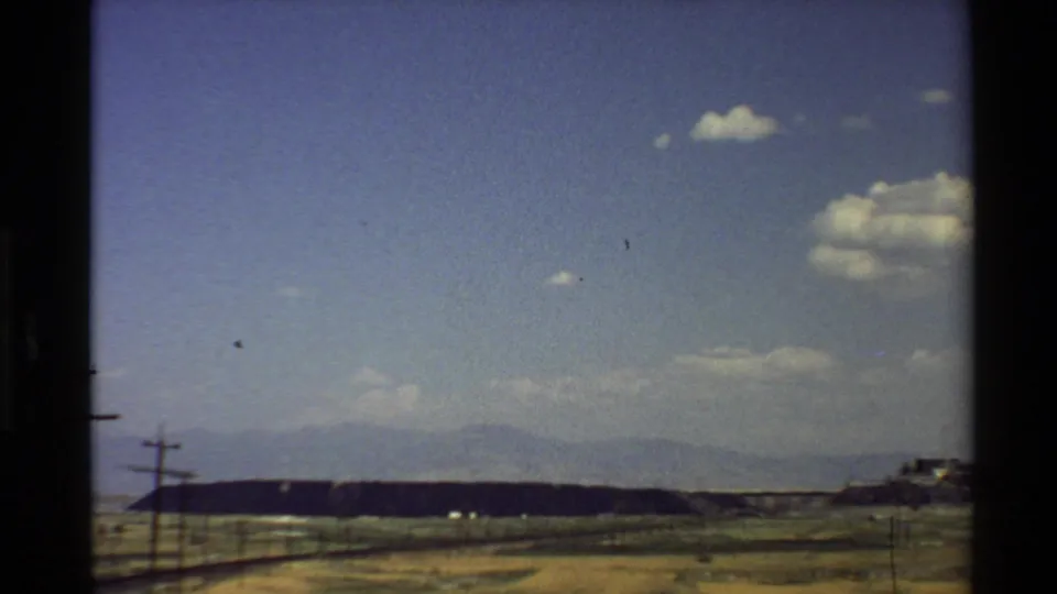 Western U.S. 8mm frame showing an open dry landscape, roadside utility poles, a low ridge, and hazy mountains beyond.