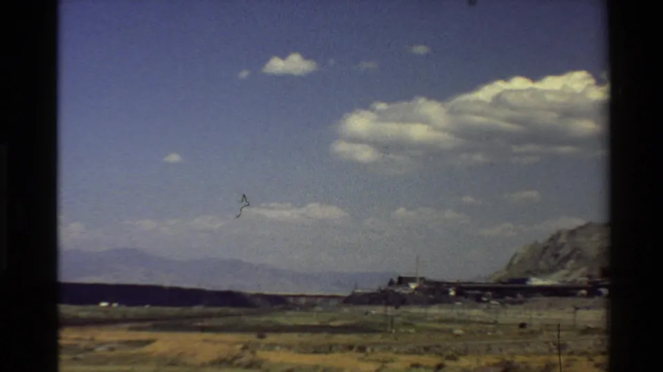 1980 roadside home-movie frame showing arid ground, distant mountains, large clouds, and an industrial facility emerging near rugged hills.