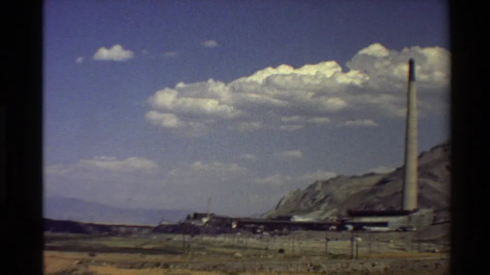 Western U.S. 8mm frame showing an industrial facility with a tall smokestack, rugged hills, dry foreground, and bright clouds.