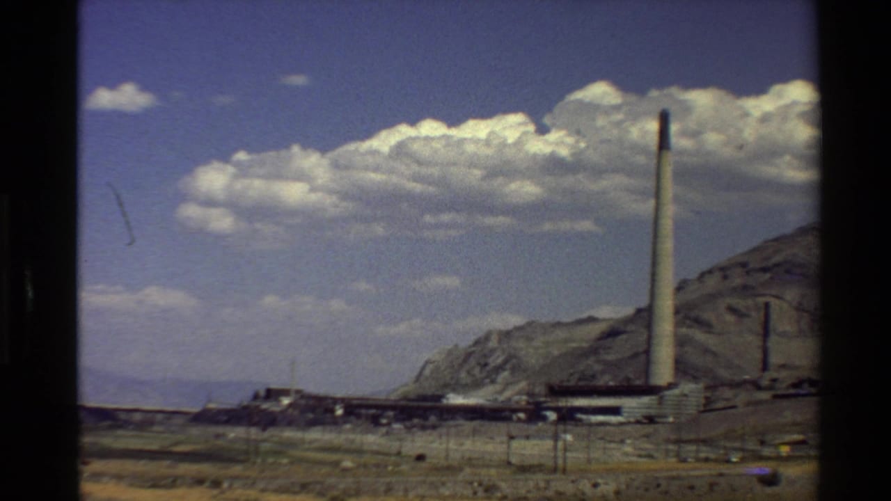 Industrial complex with a tall smokestack below mountains and clouds.