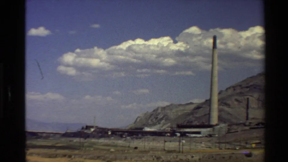 1980s 8mm view of a Western U.S. industrial complex with a tall smokestack, dry ground, mountains, and white clouds.