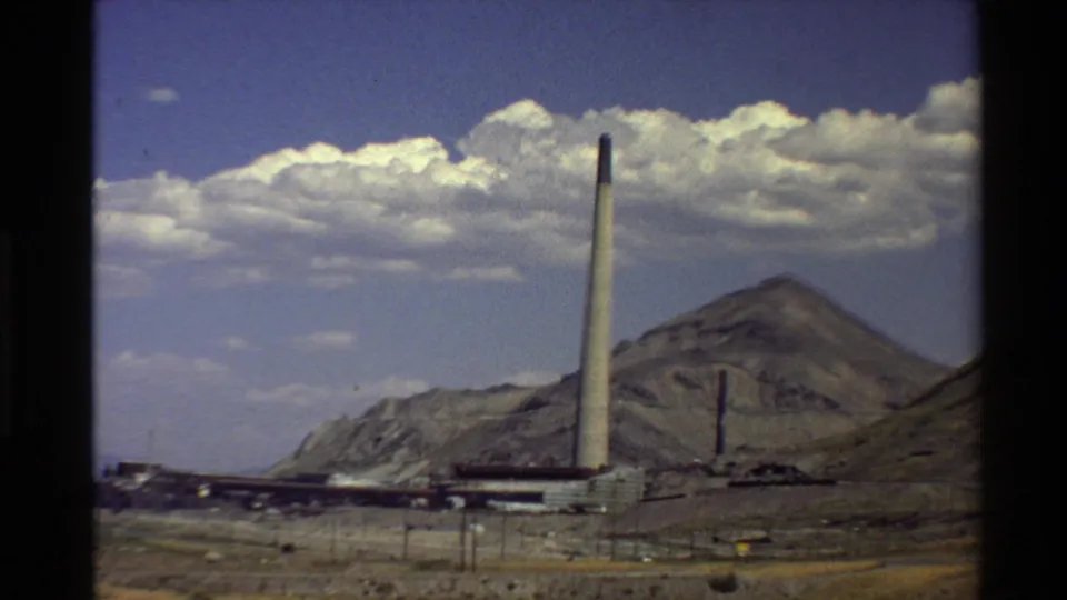 Western U.S. 8mm frame showing a factory smokestack, low industrial structures, arid terrain, and a mountain slope.