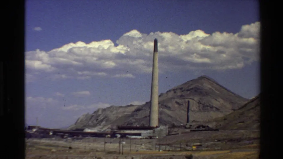 Archival 8mm frame of an industrial smokestack and factory structures set against Western U.S. mountains and a bright cloudy sky.
