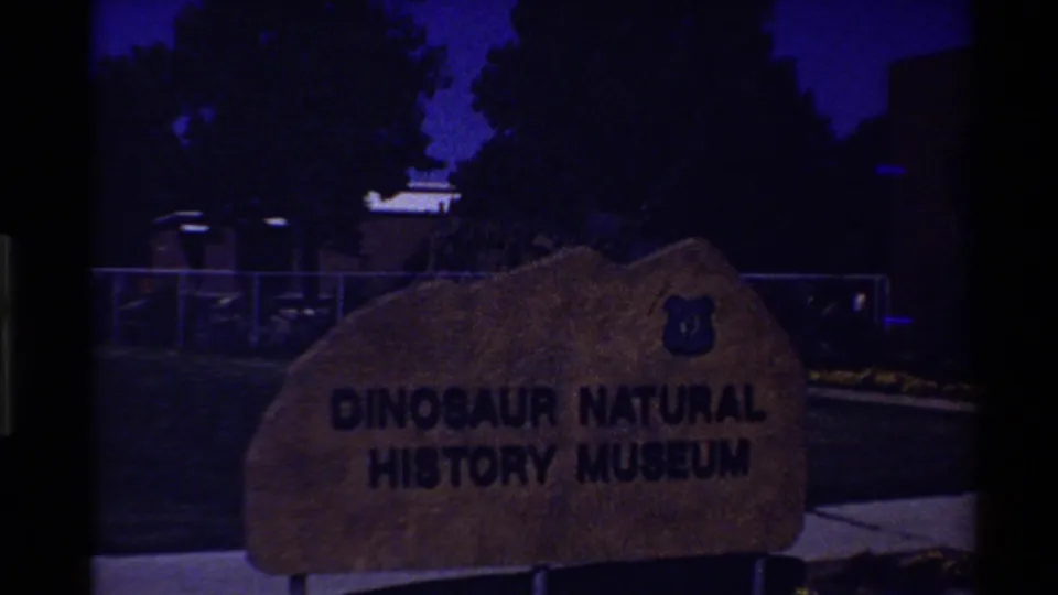 Dusk 1980 home-movie view of a stone Dinosaur Natural History Museum sign, with trees, fencing, and deep blue sky behind it.
