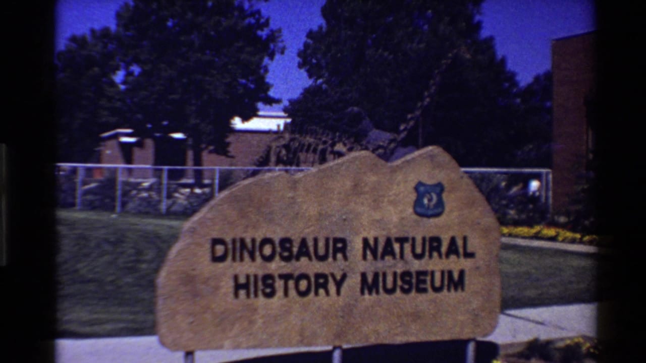 1980 8mm frame of a tan Dinosaur Natural History Museum sign outside a fenced dinosaur skeleton display in the Western USA.