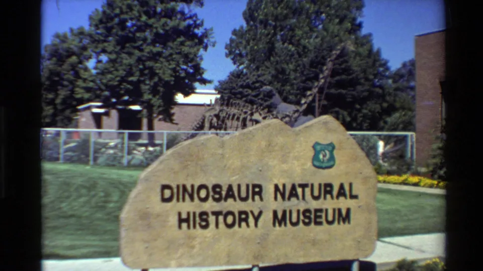 1980 home-movie frame showing the tan Dinosaur Natural History Museum sign against a green lawn and fenced outdoor exhibit.
