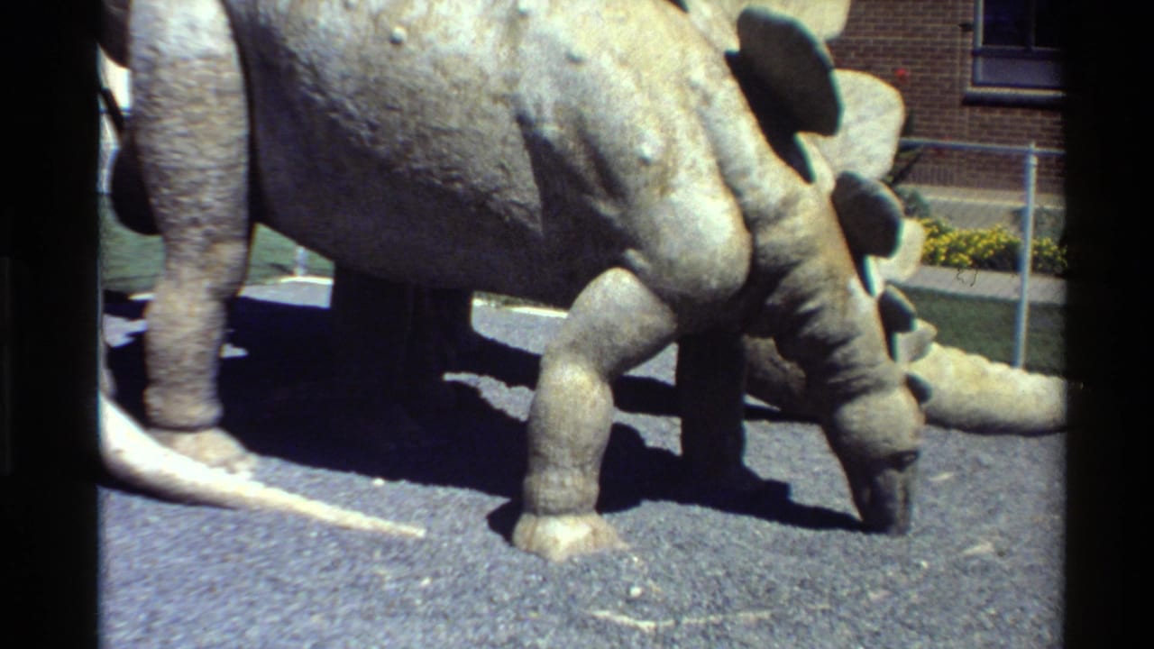 1980 Western USA 8mm frame showing a gray Stegosaurus-like museum model on asphalt with brick wall, window, and fencing behind it.