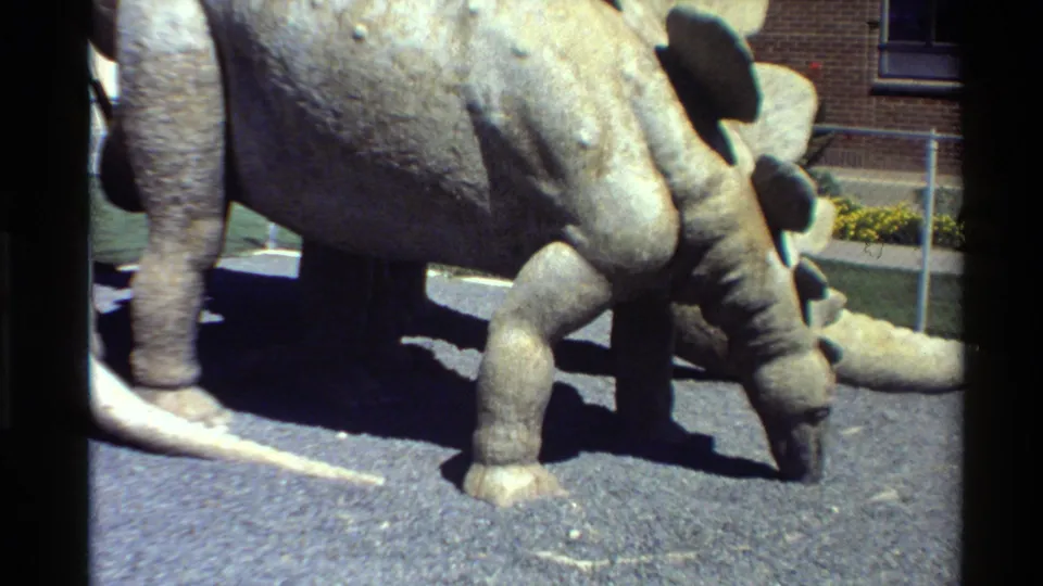 1980 Western USA 8mm frame showing a gray Stegosaurus-like museum model on asphalt with brick wall, window, and fencing behind it.