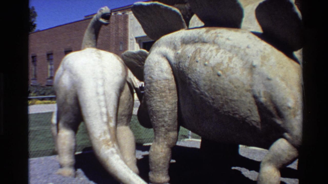 1980 Western USA 8mm frame showing faded green and white dinosaur statues near a brick building under bright sun.