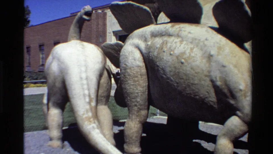 1980 Western USA 8mm frame showing faded green and white dinosaur statues near a brick building under bright sun.