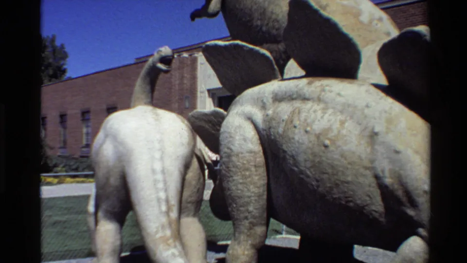 Visitor-shot 8mm view of weathered dinosaur statues, with a long-neck figure beside a larger plated sculpture.