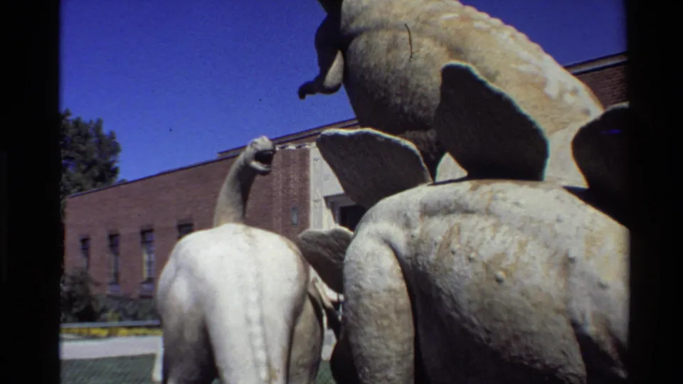 1980 home-movie frame looking upward at faded dinosaur statues in an outdoor United States display setting.