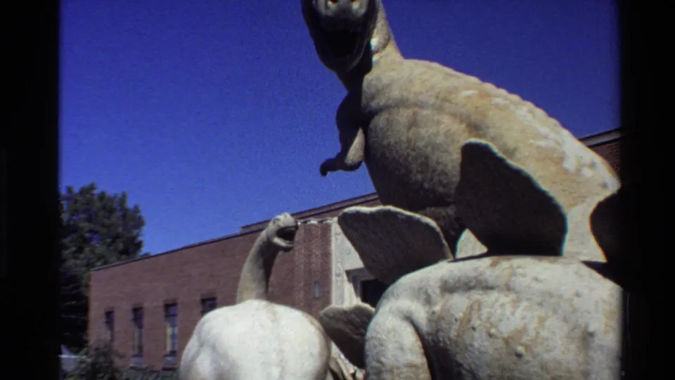 Final 8mm frame of overlapping dinosaur sculptures with weathered surfaces, blue sky, and a brick building behind them.