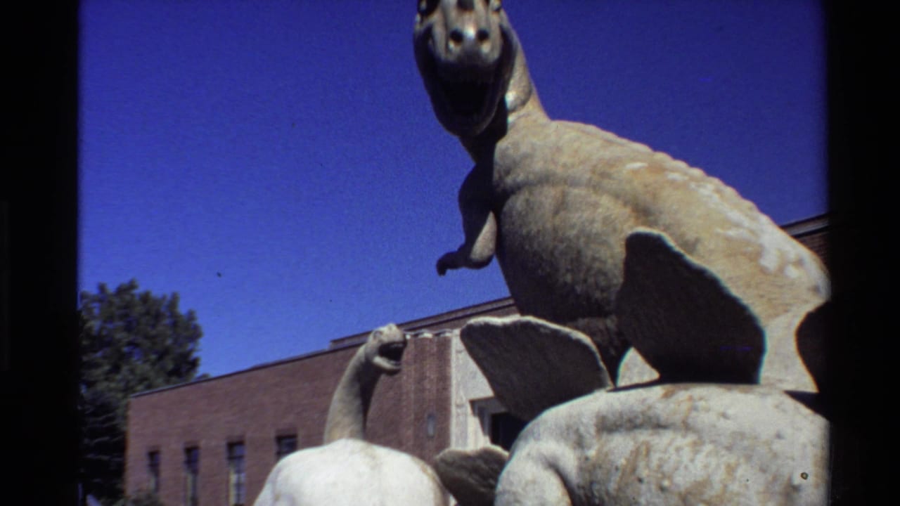 1980 Western USA 8mm frame showing a large outdoor dinosaur statue above smaller figures, with blue sky and a brick building in the background.