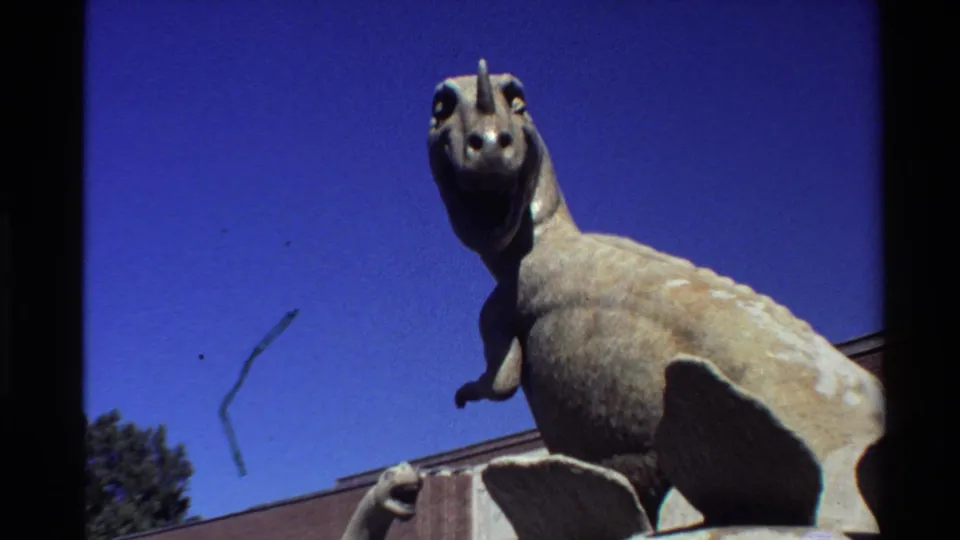 The 8mm frame moves closer to the horned dinosaur statue, with smaller statues partly visible below and a brick building at the lower edge.