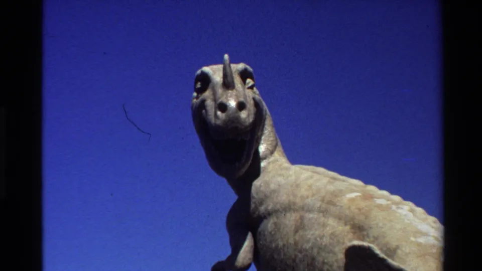 A closer 1980 home-movie view centers the dinosaur statue head and upper body, with saturated blue sky and film dust visible.
