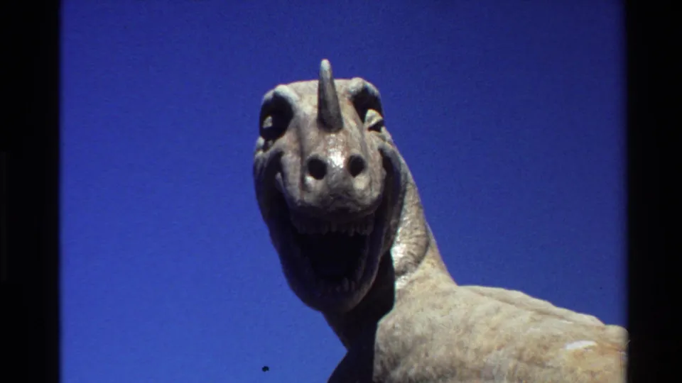 Final close 8mm frame of the outdoor dinosaur statue, emphasizing its horned face, open mouth, stone texture, and vivid sky.