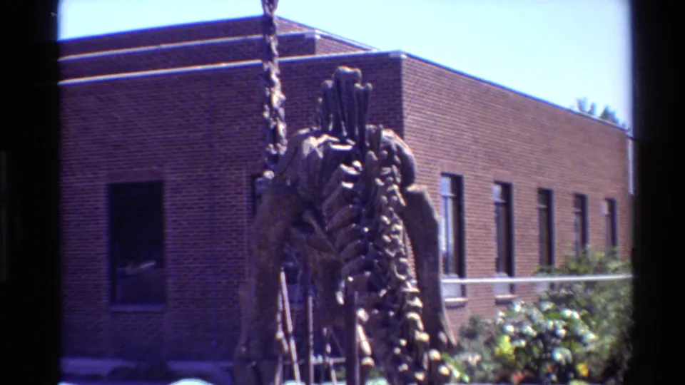 1980 Western U.S. home-movie frame showing a mounted dinosaur skeleton in front of a brick institutional building with greenery below.