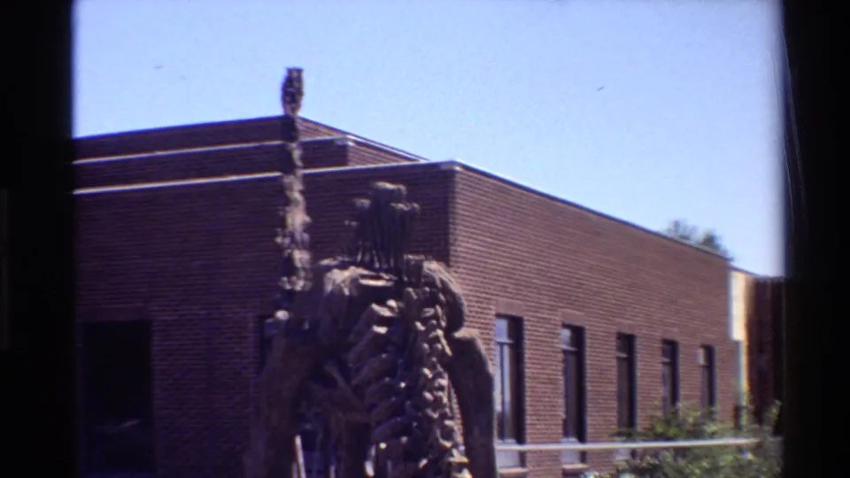 Soft 8mm frame from 1980 showing the dinosaur skeleton in front of a long brick building with windows, railing, shrubs, and sky.