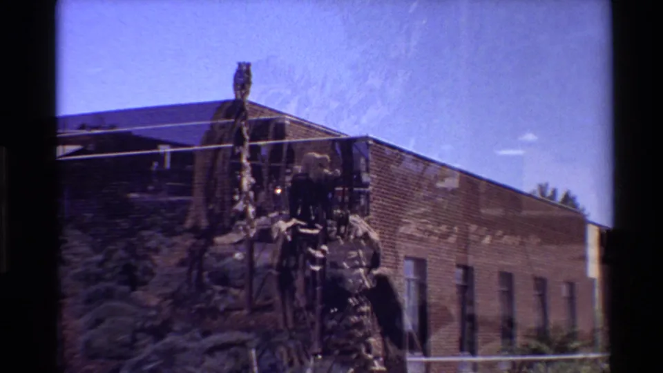 Wider 1980 Western U.S. home-movie view of the mounted dinosaur skeleton, support frame, brick building, bushes, and open sky.