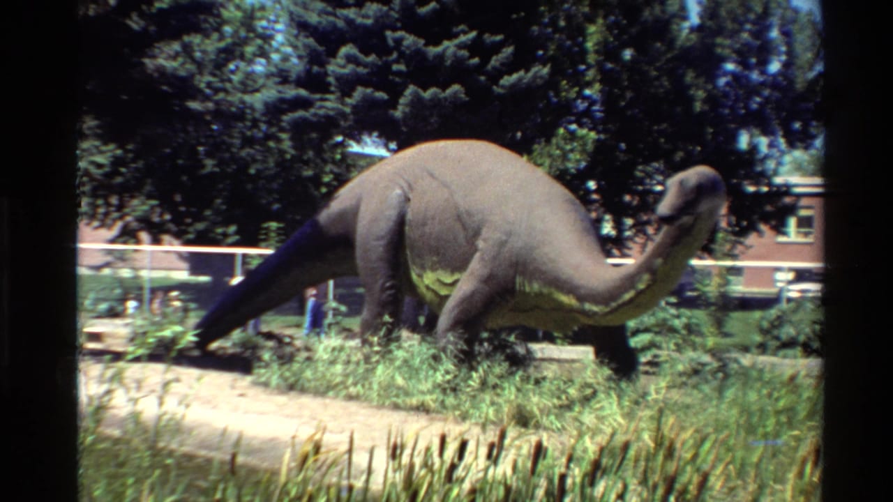 1980 United States 8mm frame showing a sauropod-like dinosaur model in a landscaped outdoor exhibit with fencing and a brick building in the background.