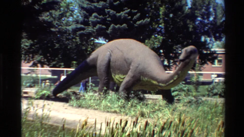 Side view of the outdoor dinosaur model, with its long neck and tail visible against grass, trees, a white fence, and a red brick building.