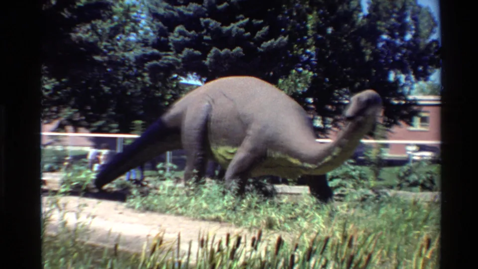 Final frame holds on the outdoor dinosaur model amid grass and trees, with a white fence and red brick building still visible behind the exhibit.