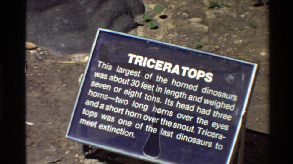 Late-frame 8mm close-up of the blue Triceratops placard outdoors, showing white lettering, dirt ground, rocks, and film-frame borders.