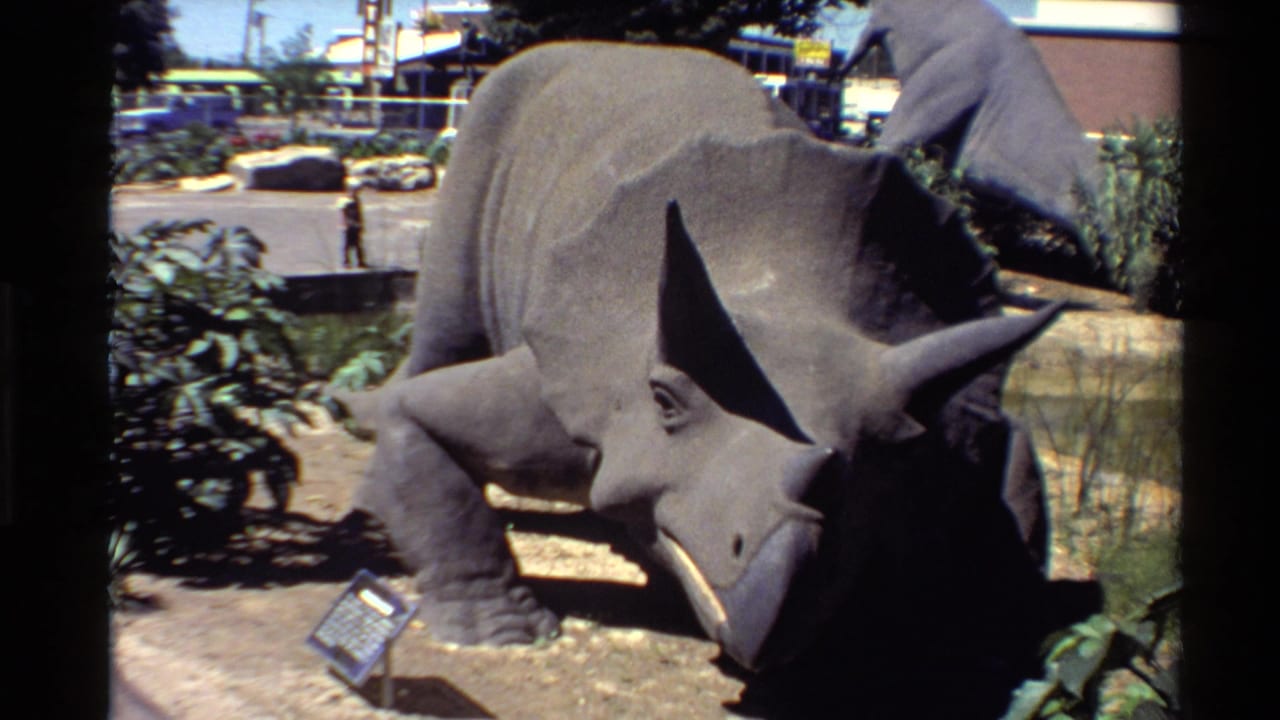 1980 U.S. 8mm frame showing a gray outdoor triceratops model with horns, frill, dirt display bed, plants, and a small sign.