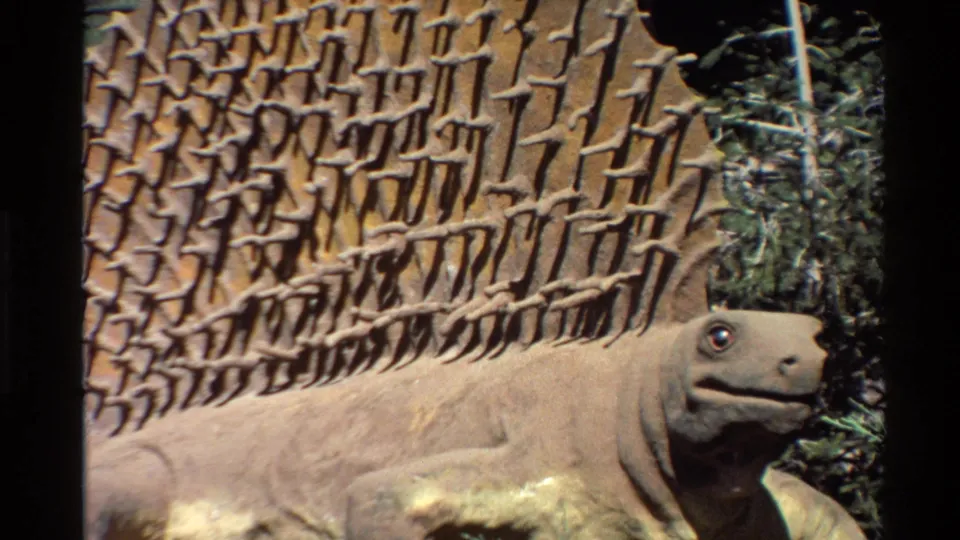 1980 United States 8mm frame showing a wider view of the Dimetrodon-like statue, its spined back, and surrounding artificial foliage.
