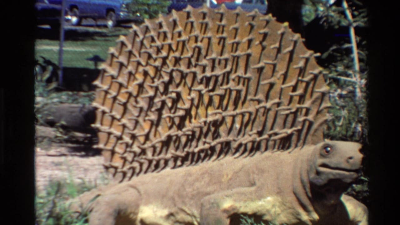 1980 Western U.S. home-movie frame showing a brown Stegosaurus statue in planted grounds with parked cars beyond a chain-link fence.