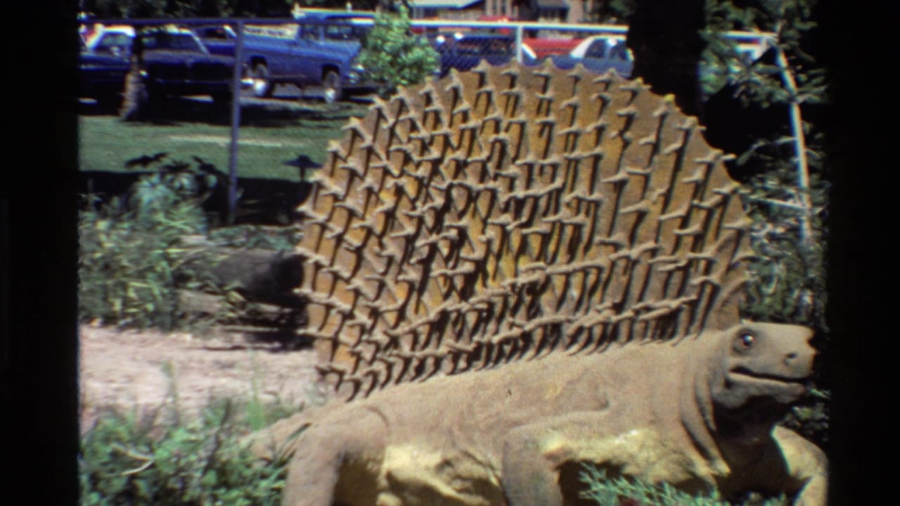 Stegosaurus statue partly framed by foliage with cars and fence in back.